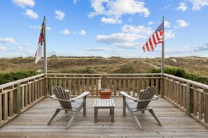 A wooden deck with two Adirondack chairs and a small table, overlooking a beach with dunes and flags.