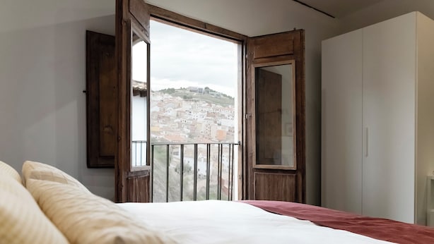 A bedroom with a bed, a wardrobe, and a balcony with a view of a town. - La Casa del Orfebre (Cuenca)