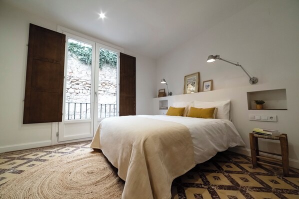 Un dormitorio con una cama grande, una lámpara de pie, una mesita pequeña y vista a una pared de piedra a través de una ventana.