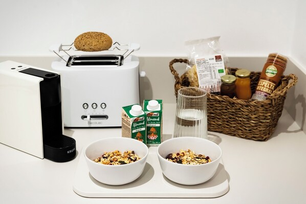 A kitchen counter with a toaster, two bowls of granola, a carton of almond milk, a glass of water, and a basket containing bread and jars.