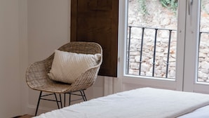 A wicker chair with a cushion in a room with a bed and a view of a stone wall and greenery outside. - La Casa del Orfebre (Cuenca)