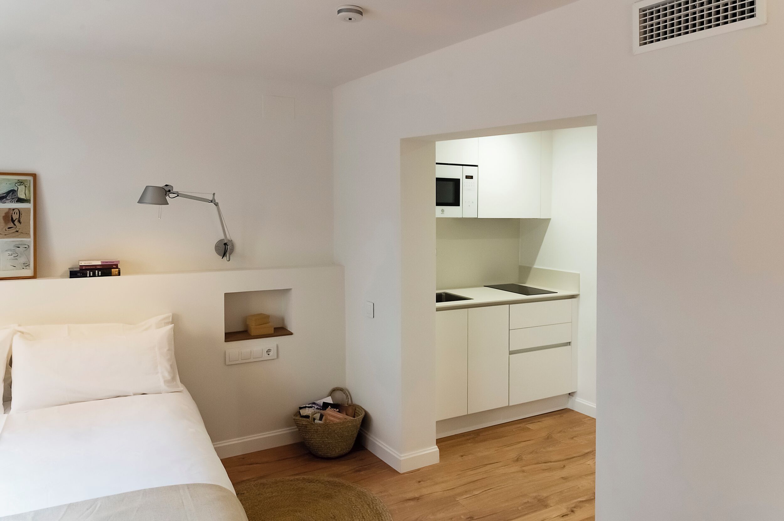 Habitació petita amb llit, escriptori, làmpada, prestatge petit i cuineta.