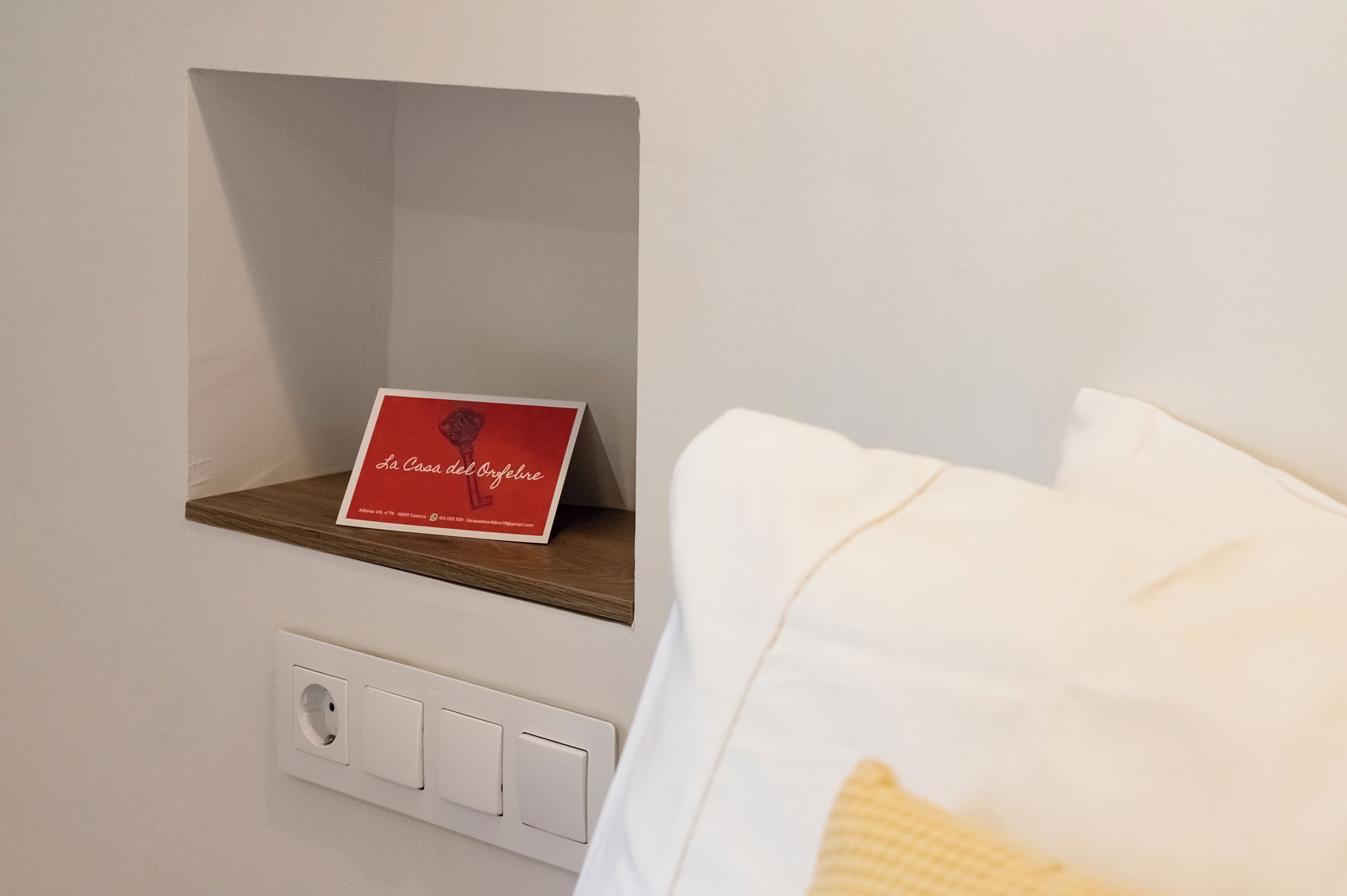 Een hotelkamer met een houten plank waarop een rood bord staat, een wit bed en een stopcontact.