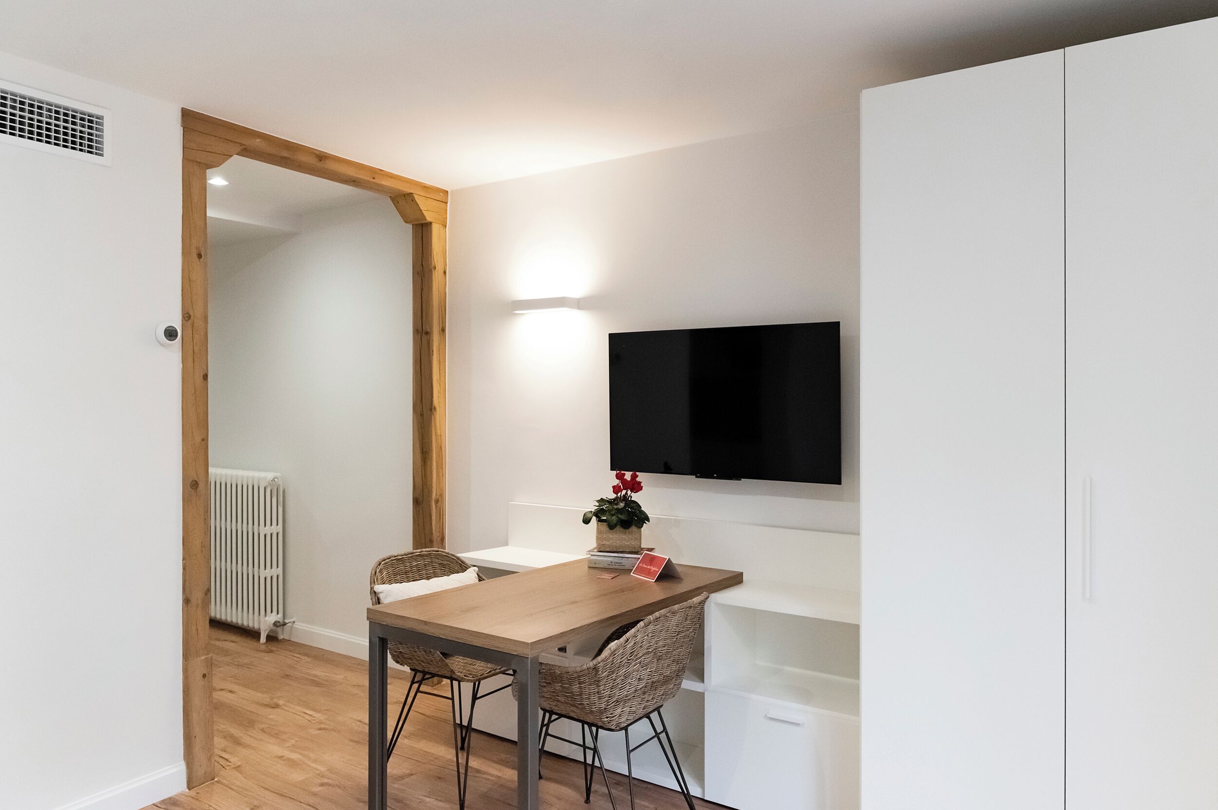 A modern kitchen with white cabinetry, a wooden dining table, wicker chairs, and a flat-screen TV mounted above the counter.