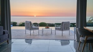 Una habitación moderna con vistas a la puesta de sol sobre el mar, que incluye una zona de estar con dos sillones y una mesa, además de un comedor con mesa y sillas.
