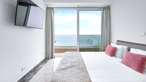 Habitación de hotel con una cama grande, un televisor de pantalla plana montado en la pared y vista al mar a través de una puerta corrediza de vidrio.