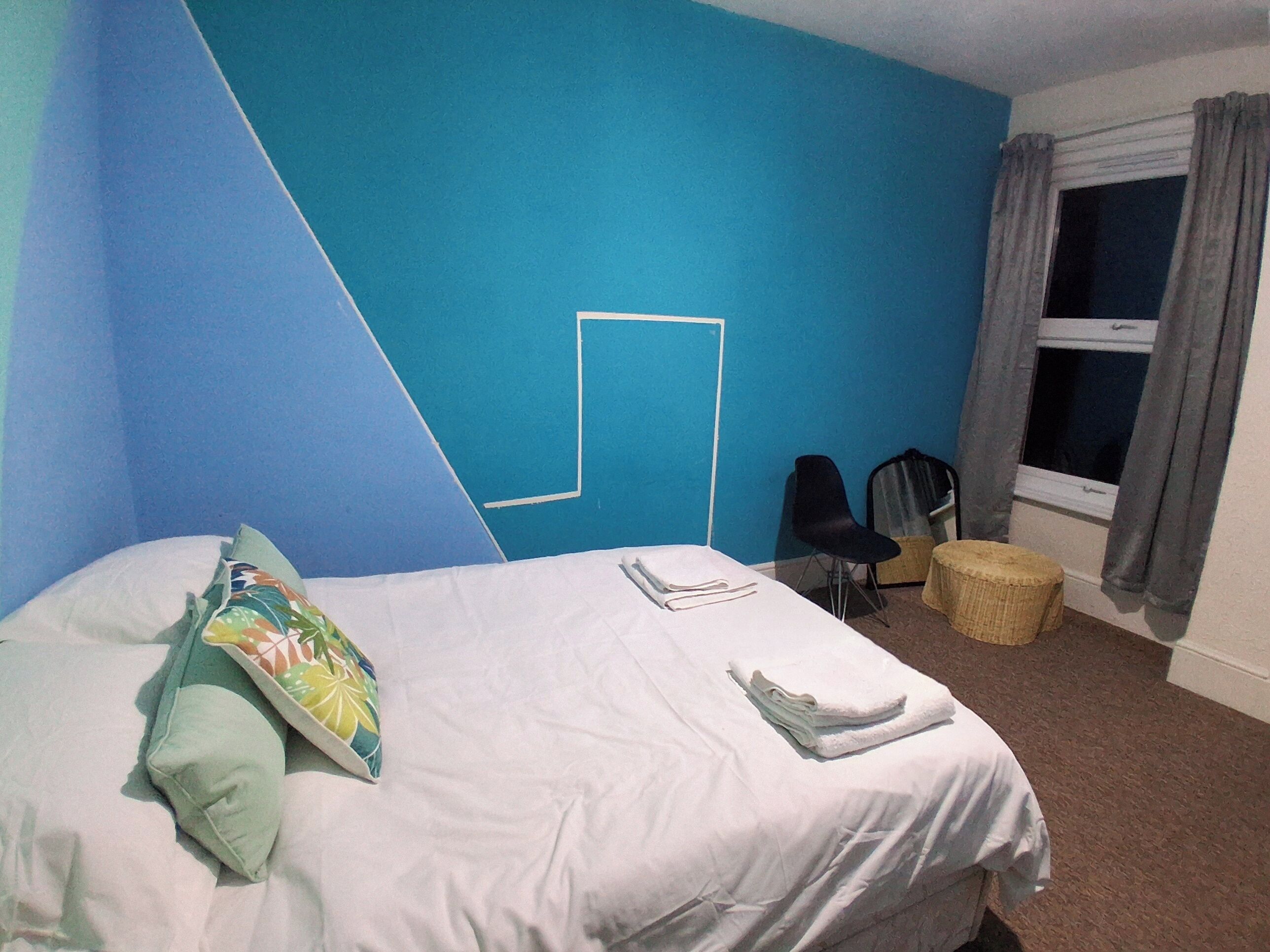 A hotel room with a bed, a chair, a small stool, a window with curtains, and a blue wall with a white line design.