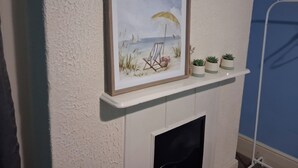 Un tableau représentant une scène de plage, encadré, est accroché au-dessus d’une cheminée blanche, et trois petites plantes en pot sont disposées en dessous.