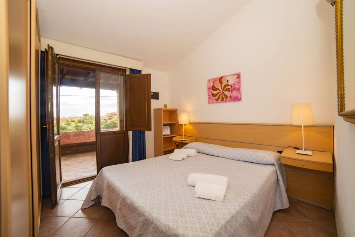 A hotel room with a bed, two bedside tables, a nightstand, a painting on the wall, and a sliding door leading to an outdoor area.