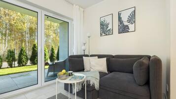 Un salon moderne avec un canapé gris, une petite table ronde sur laquelle repose un magazine, et une grande porte-fenêtre coulissante donnant sur un jardin.