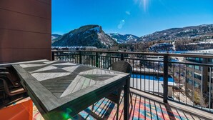 A balcony with a wooden table and chairs, overlooking a snowy mountain landscape and a pool.