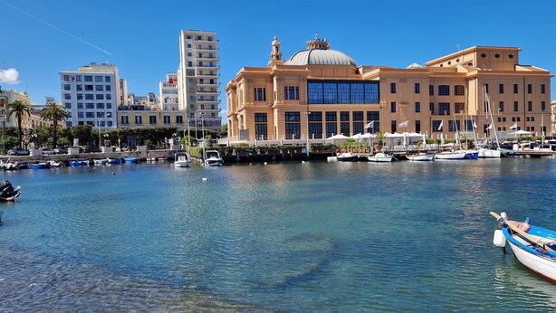 Una marina con imbarcazioni ormeggiate, un grande edificio con cupola e colonne, e una gru edile sullo sfondo.