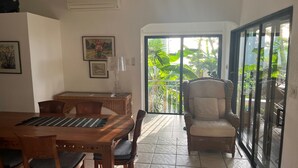 Zona pranzo con tavolo e sedie di legno, una sedia in vimini e una porta vetrata che dà su un balcone con piante tropicali.