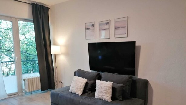 A living room with a grey sofa, two white fluffy cushions, a large flat-screen TV mounted on the wall, and a floor lamp. - Quiet apartment 4 min from the cathedral with office and balcony (Köln)