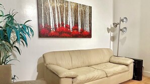 Un salon avec un canapé beige, un tableau représentant une forêt, un lampadaire et une plante en pot.