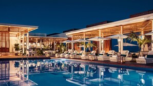 Uma área à beira da piscina com espreguiçadeiras e guarda-sóis, uma piscina cristalina e um edifício com espaço de estar coberto.