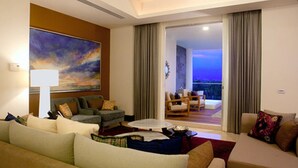 Uma sala de estar com sofá, poltronas, mesa de centro e um quadro na parede. Há uma vista do exterior através de uma porta de vidro.
