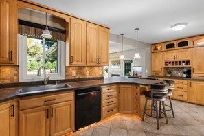 A kitchen with wooden cabinets, a dishwasher, a large island with bar stools, pendant lighting, and a window with a view of greenery.