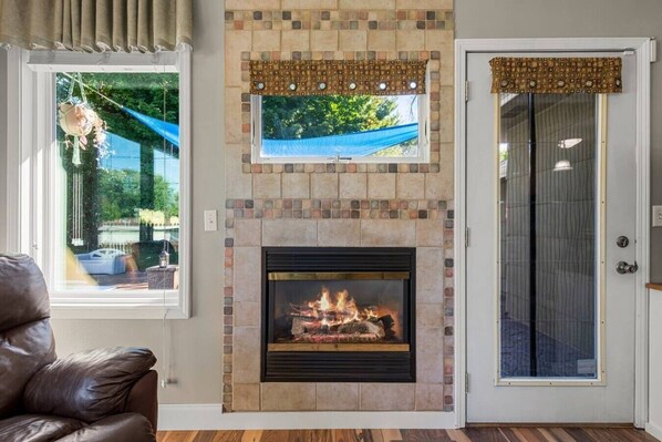 A fireplace with a stone surround, a window with a view of greenery, and a door with a patterned curtain.