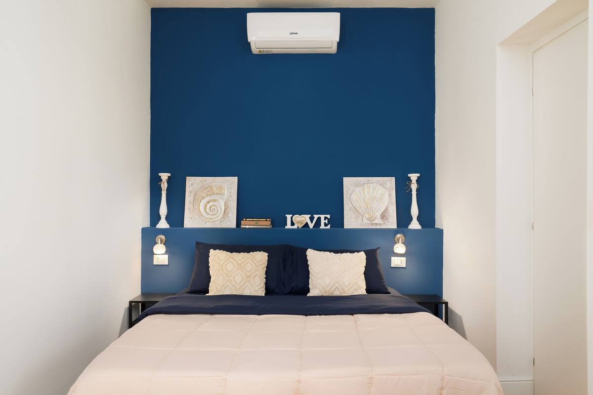 A bedroom with a blue accent wall, a bed with a beige and navy duvet, two bedside lamps, and a wall-mounted air conditioner.