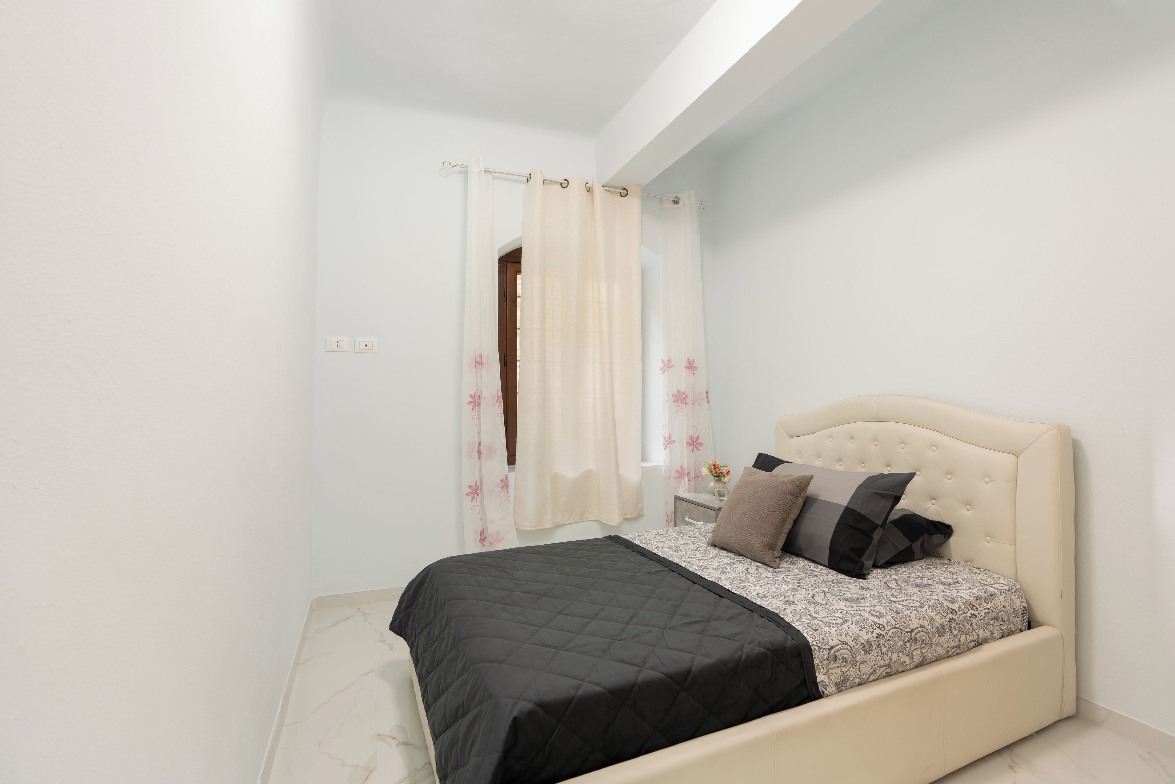 Un dormitorio con cama, mesita de noche, ventana con cortinas y puerta.