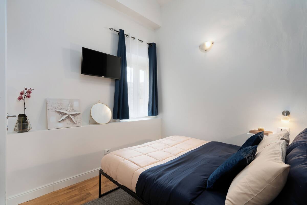 A bedroom with a bed, a TV mounted on the wall, a window with curtains, a bedside table with a lamp, and a wall-mounted light fixture.