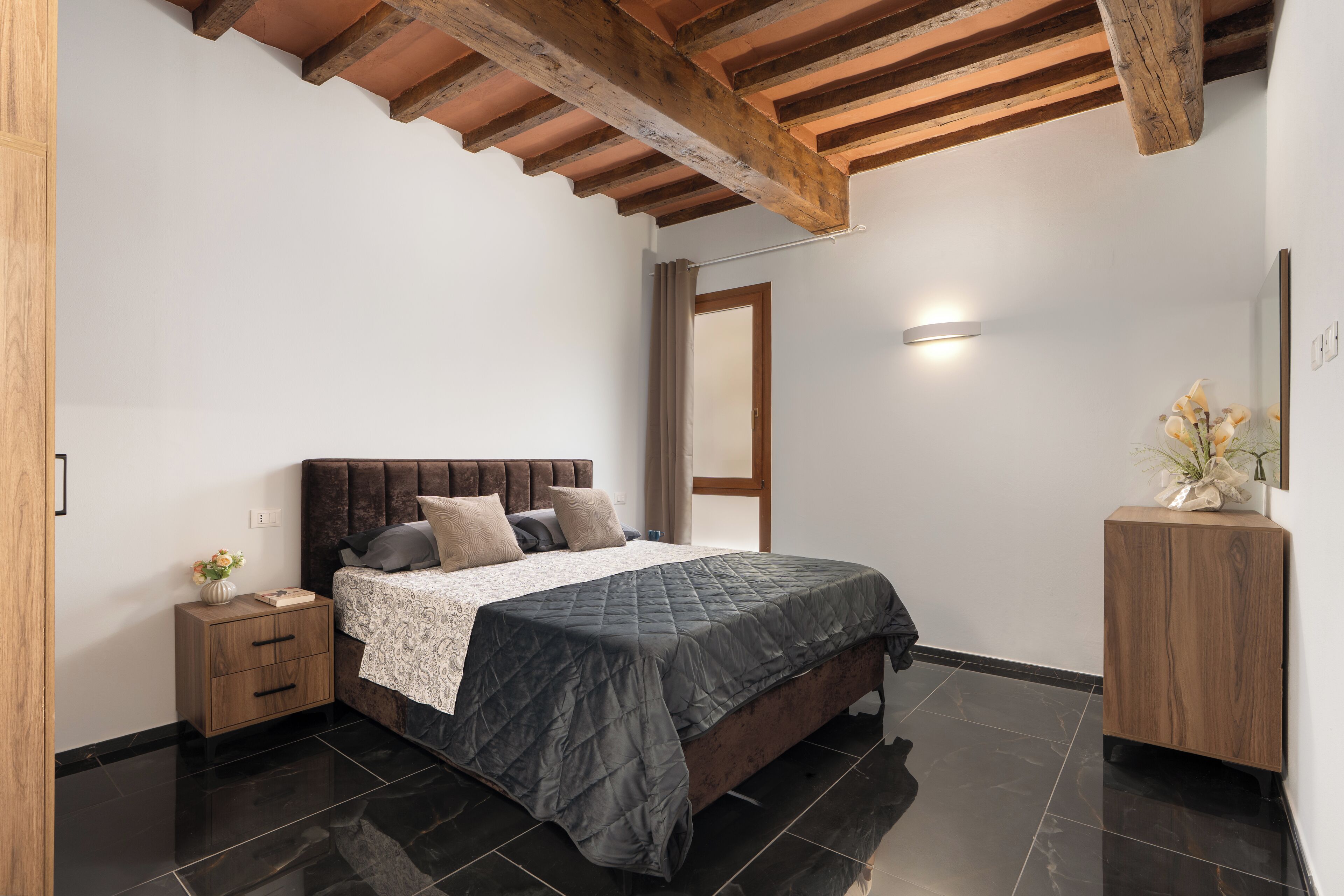 Una camera da letto con una testiera in legno, un comodino con cassetti, una coperta trapuntata scura e un tavolino da notte con un vaso di fiori.