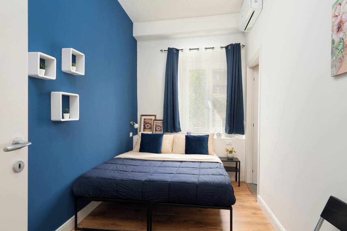 A bedroom with a bed, a window with curtains, a bedside table, and wall-mounted shelves with plants.