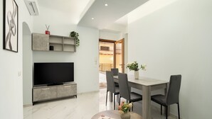 Un soggiorno moderno con una TV a schermo piatto, mobili contenitori grigi, un tavolo da pranzo con sedie e un piccolo tavolino rotondo con un libro e dei fiori.