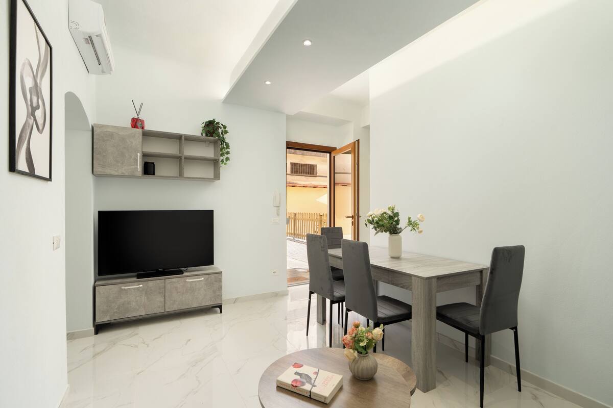 A modern living room with a flat-screen TV, grey cabinets, a dining table with chairs, and a small round table with a book and flowers.