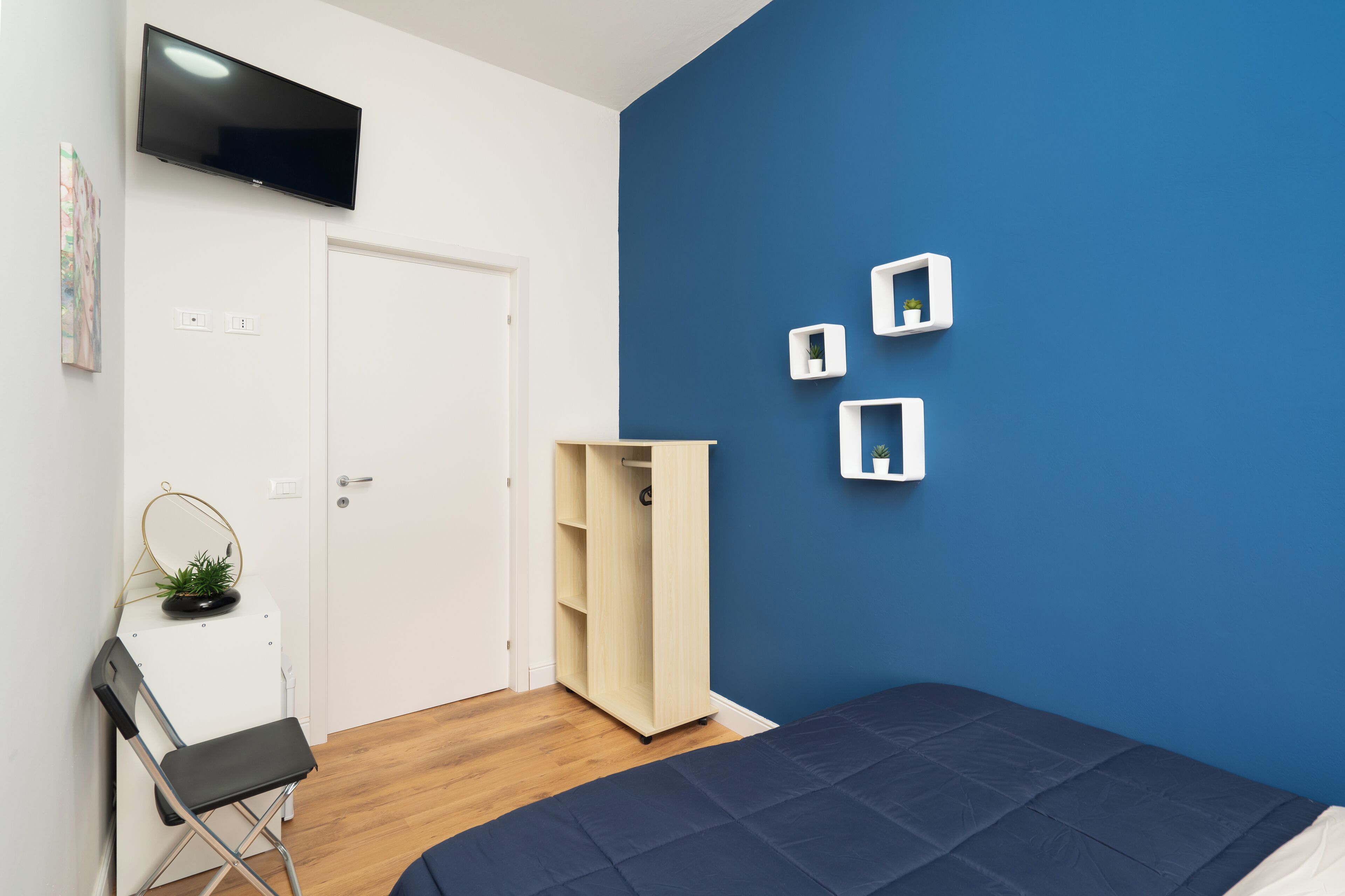 Un dormitorio con una pared azul, una cama, un televisor montado en la pared y un armario.