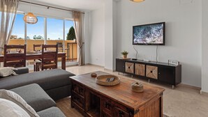 A modern living room with a sofa, coffee table, and TV. There's a balcony with a dining area and a view of palm trees. - Casa Bonita de La Cala (Mijas)