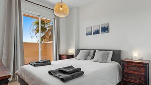 A bedroom with a large bed, two bedside tables, a hanging light fixture, and a view of palm trees through a sliding door. - Casa Bonita de La Cala (Mijas)