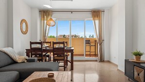 A modern living room with a sofa, a dining table, and chairs. There is a balcony with a view of the sky and buildings. - Casa Bonita de La Cala (Mijas)