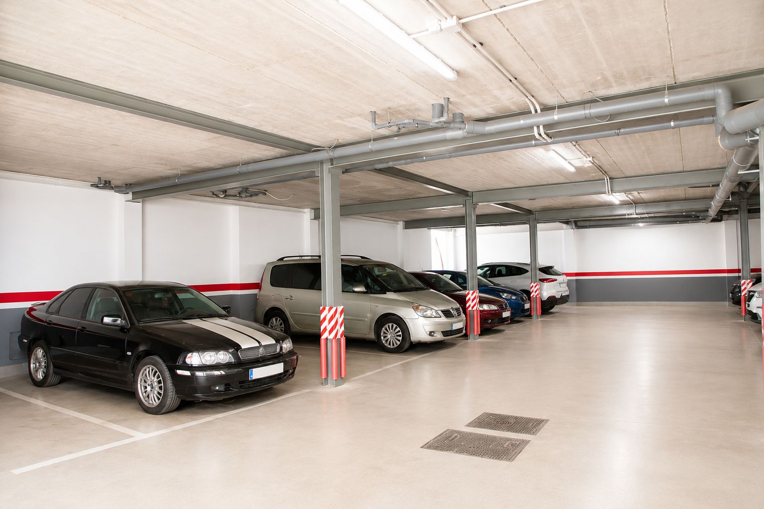 Un parking à plusieurs niveaux où plusieurs voitures sont garées sous un plafond en béton, avec des tuyaux apparents.