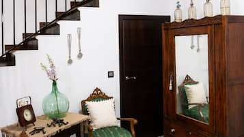 Una escalera con barandilla de madera y balaustres negros, un armario de madera con espejo, una silla tapizada de verde, una mesita con un reloj y un jarrón, y un suelo estampado.
