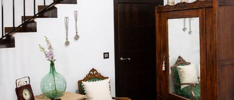 A staircase with a wooden handrail and black balusters, a wooden cabinet with a mirror, a green upholstered chair, a small table with a clock and a vase, and a patterned floor.
