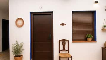 Entrada de hotel con puerta de madera, planta en maceta, silla y ventana con persianas.