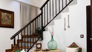 Un escalier doté d’une rampe en bois et de balustres noires, menant à un palier où se trouvent deux fauteuils recouverts de tissu vert, une petite table avec un vase et une horloge murale.