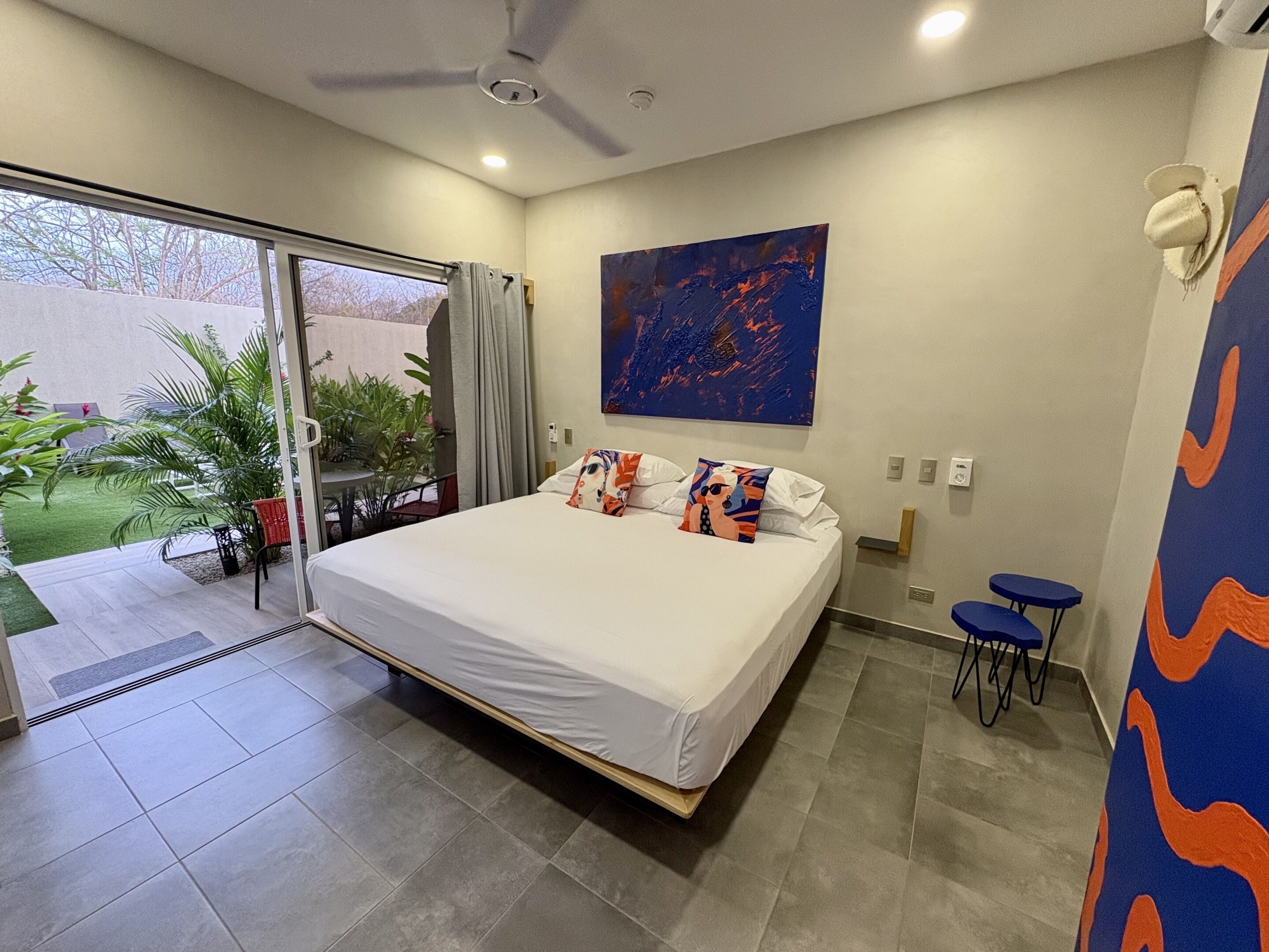 Un dormitorio con cama, ventilador de techo, un cuadro y vistas a un jardín por una puerta corredera.
