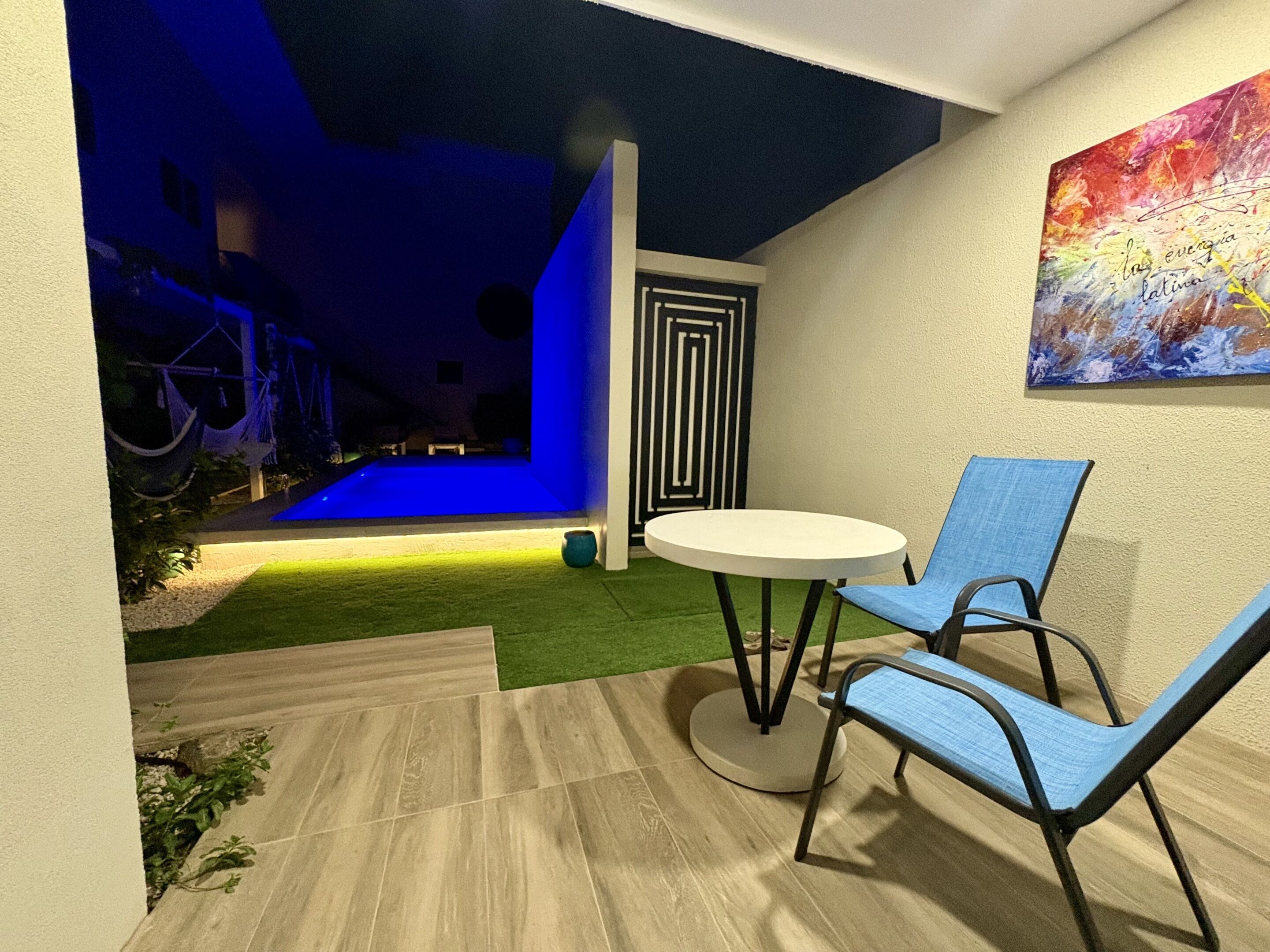 A modern outdoor patio with a round table and two blue chairs, a small garden area with artificial grass, and a colorful abstract painting on the wall.