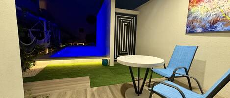 A modern outdoor patio with a round table and two blue chairs, a small garden area with artificial grass, and a colorful abstract painting on the wall.