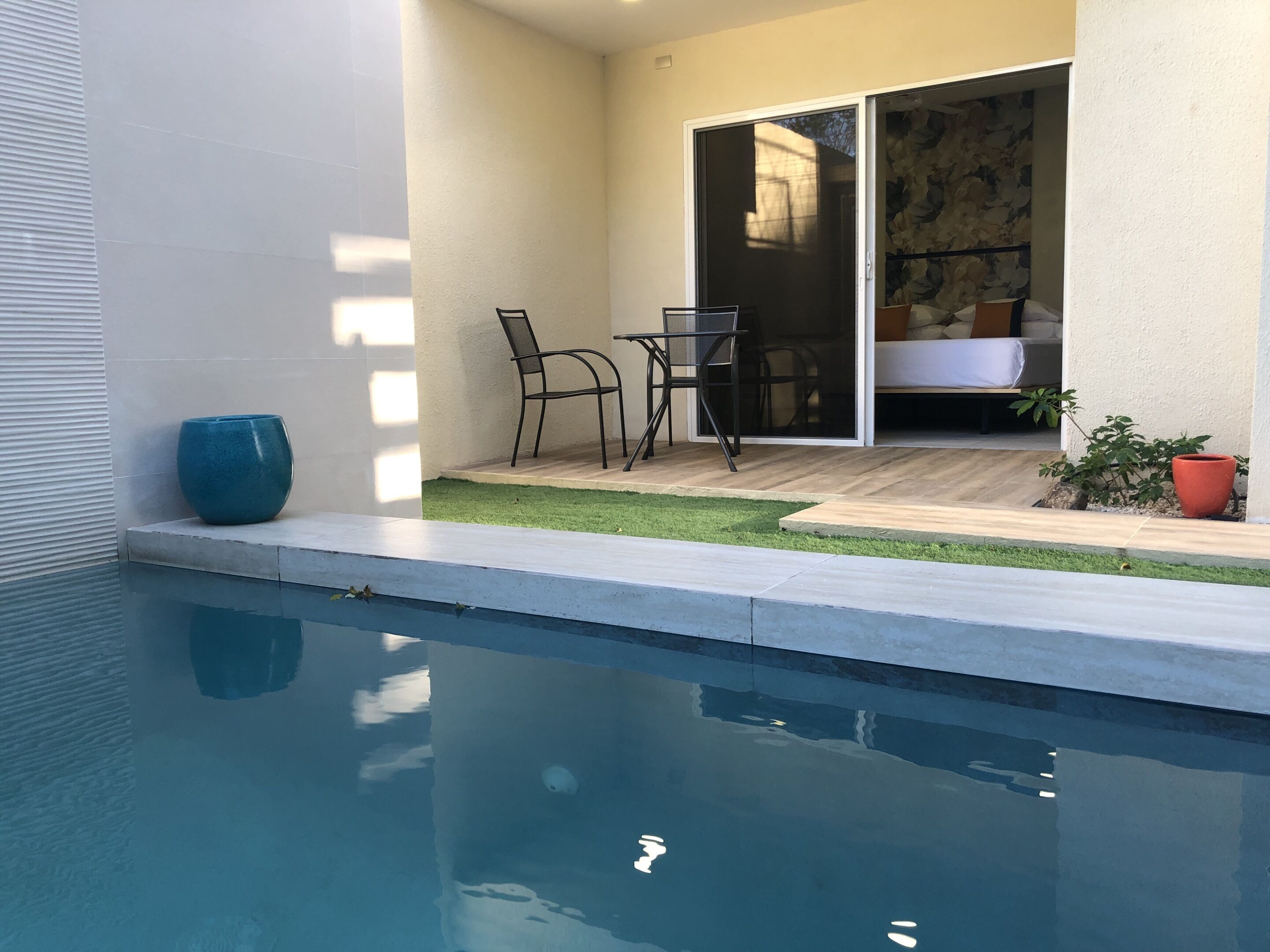 En pool med ett uteområde, en skjutdörr i glas som leder in till ett sovrum och en liten trädgård med en krukväxt.