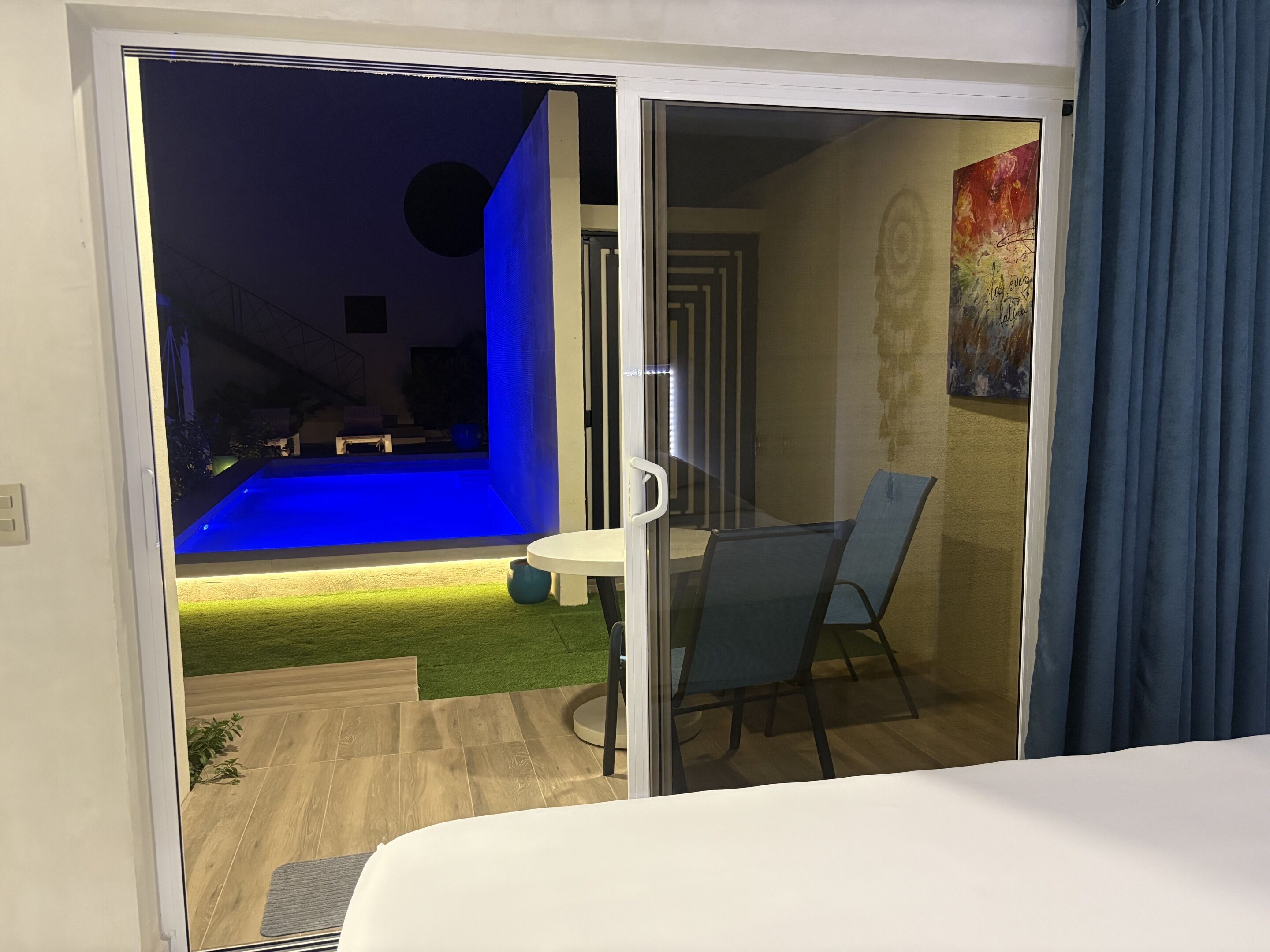 A hotel room with a sliding glass door leading to a balcony with a blue-lit pool and a small table with chairs.
