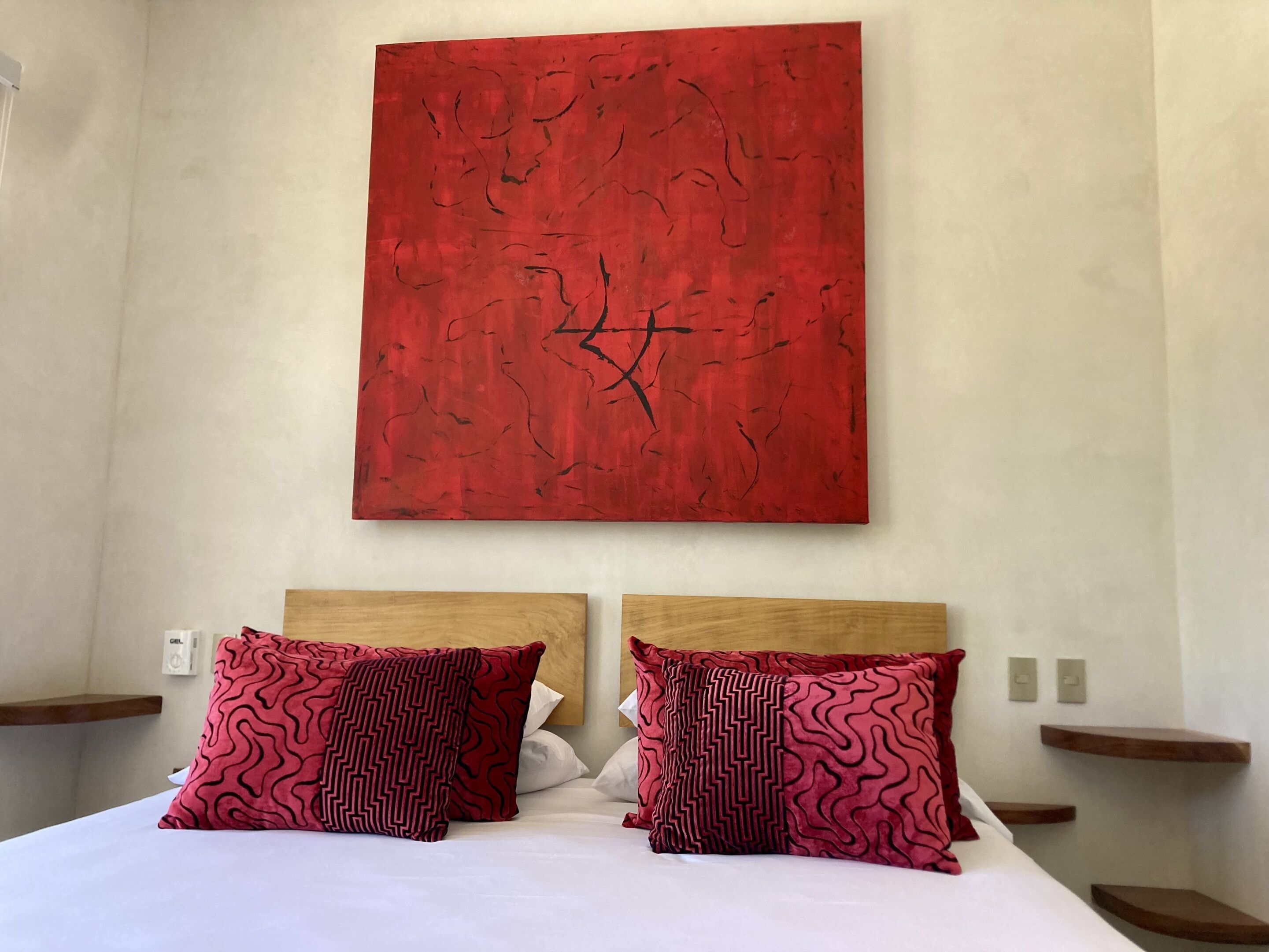 Une chambre d’hôtel avec un grand tableau abstrait rouge au mur, deux têtes de lit en bois et des coussins assortis à motifs.