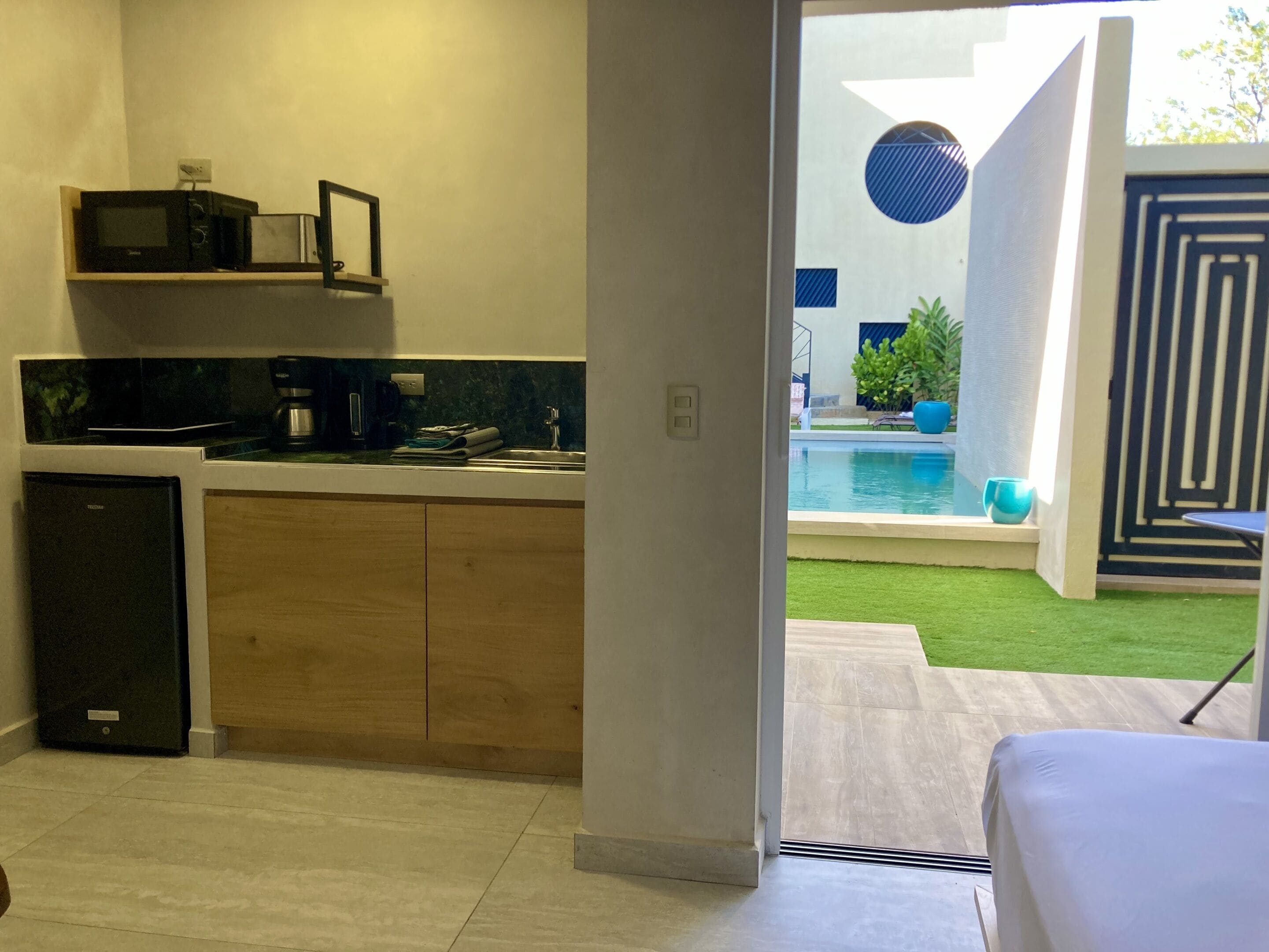 Una cocina moderna con microondas integrado, cafetera y lavabo. Un refrigerador pequeño se encuentra a la izquierda. La habitación tiene un ventanal amplio que da a una zona de piscina con un adorno circular azul y una planta en maceta.