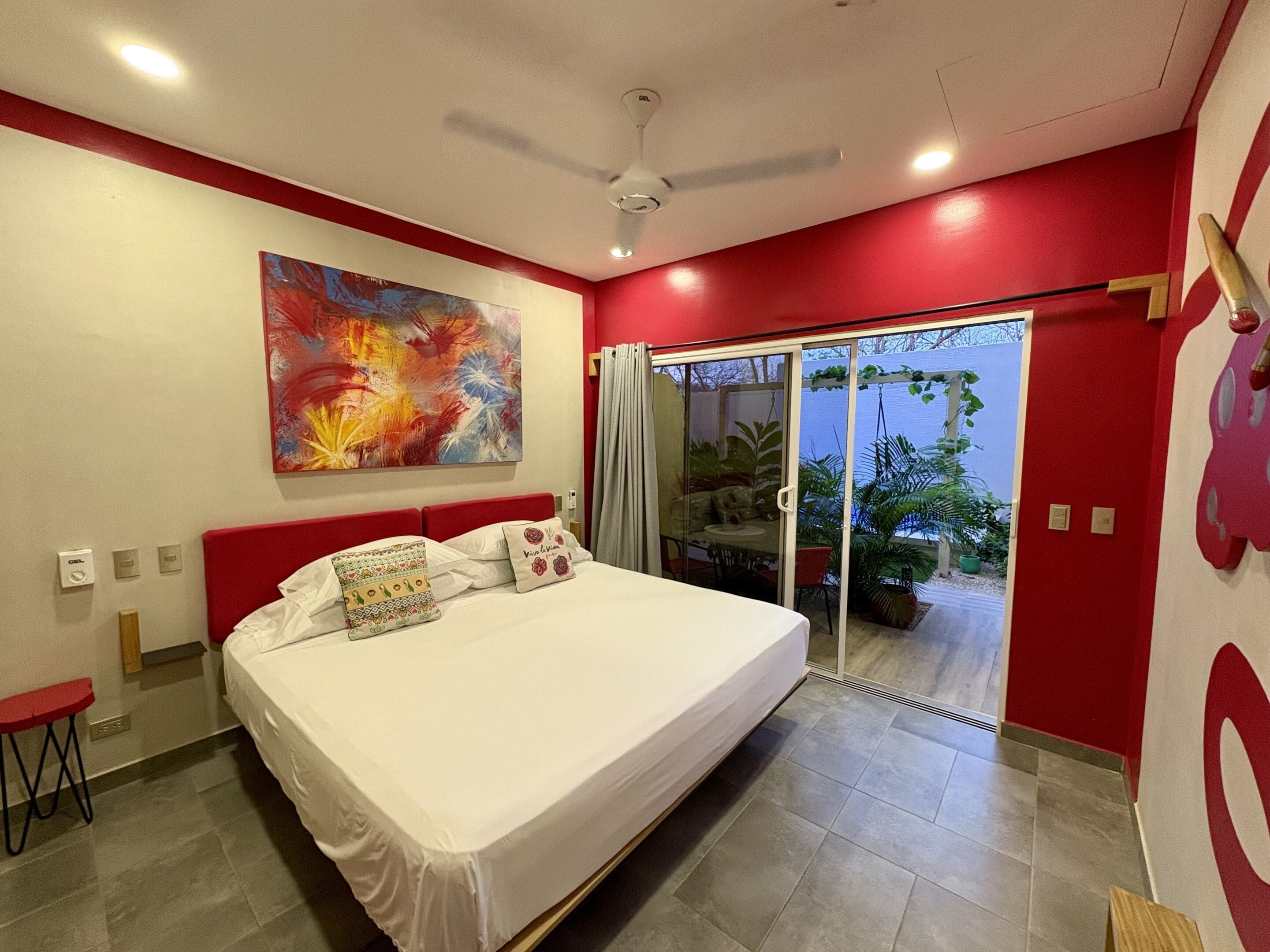 A bedroom with a bed, a ceiling fan, a large abstract painting, and a sliding glass door leading to a balcony with plants.