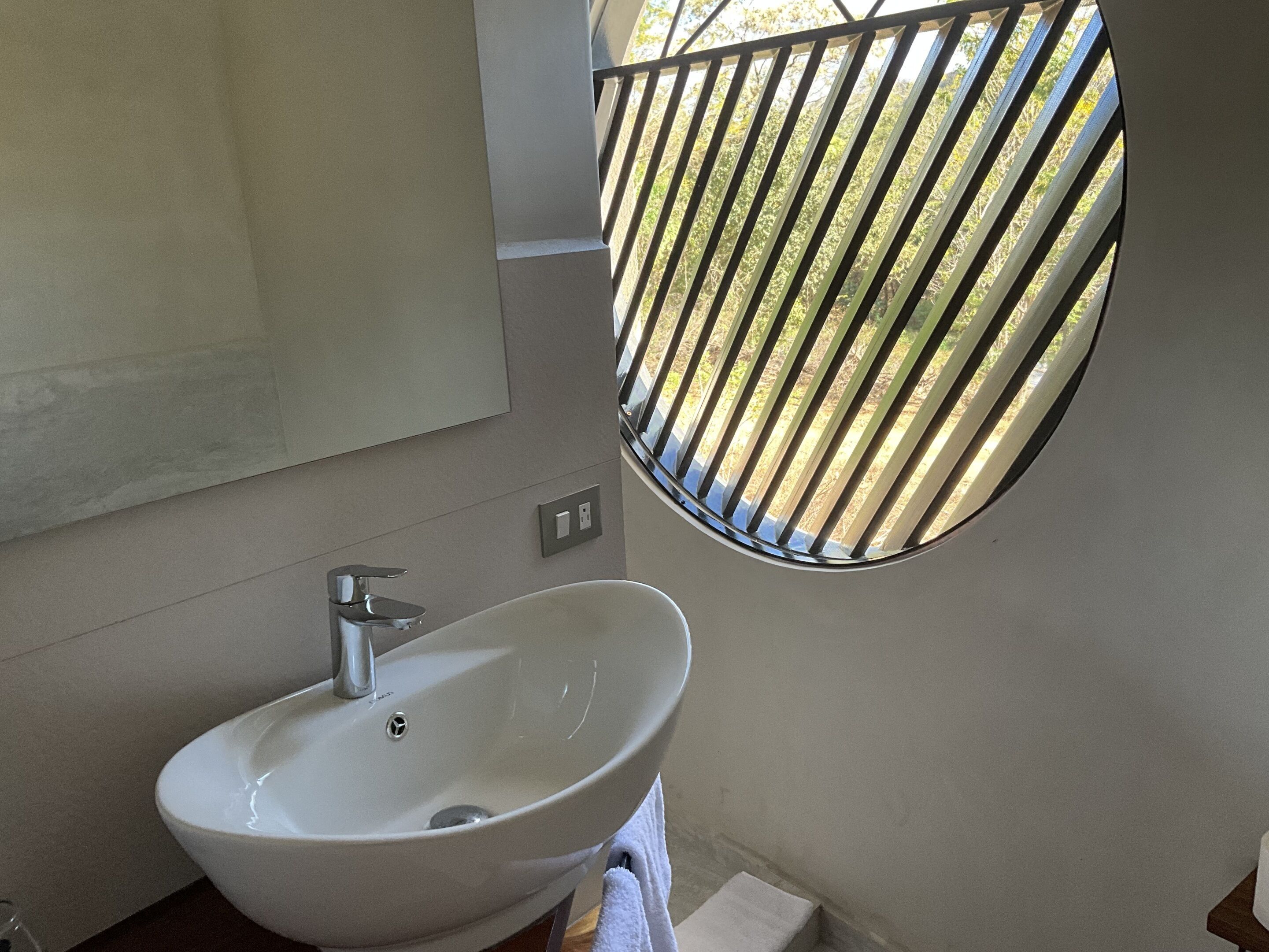 Une salle de bain avec un lavabo blanc, un miroir et une fenêtre ornée d’une grille métallique.