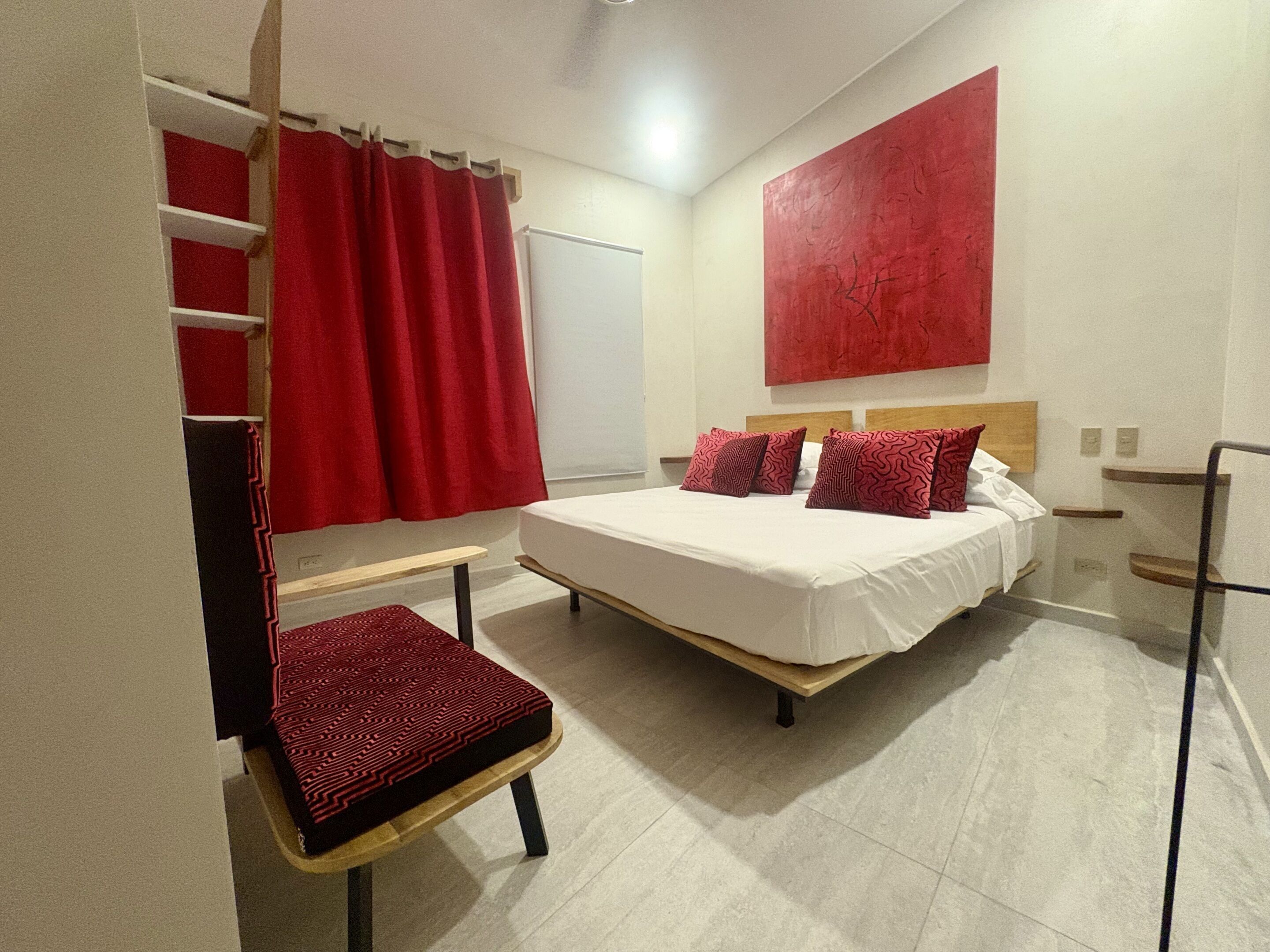 Un dormitorio con cama, una silla, una cortina roja y un cuadro abstracto rojo en la pared.