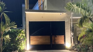 Ingresso di un edificio moderno con una porta scura, un vialetto in cemento e vegetazione circostante.